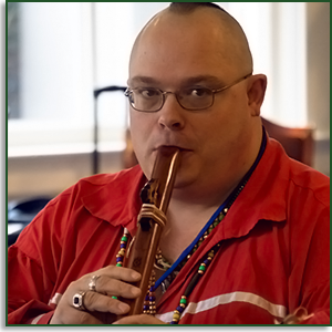 A man looks at the camera while he plays a flute. He wears indigenous necklaces and rings.
